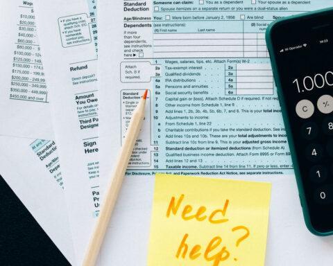 photo of tax forms spread out on a table with a sharpened pencil and calculator on top of the paperwork. A sticky note on the top sheet has the question, "Need Help?"