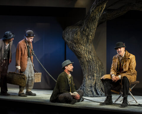A scene from the play, "Waiting for Godot," with two men standing, one man kneeling, and one man sitting on a camp stool. One of the men standing has a leash around his neck.