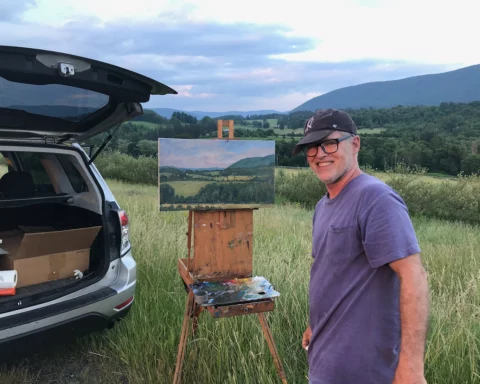 Photo of a middle-aged man wearing a tee-shirt and baseball cap standing in front of an easel at the top a hill of hayfield and a small apple orchard, with a sweeping vista in the background.