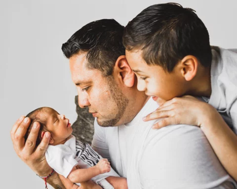 A young father gazes at the newborn baby he cradles in his hands as a young boy looks over his shoulder.