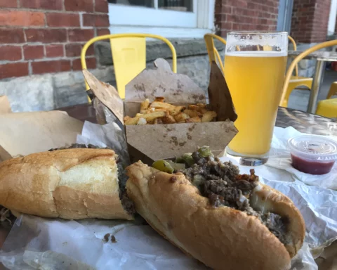 Close-up photo of a Philly Cheese Steak sandwich in the foreground with a cardboard box of French fries and a glass of beer in the background.