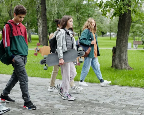 A photo of a group of male and female high school aged students walk together along a sidewalk in a grassy, parklike setting with trees in spring or summer, some carrying skateboards.