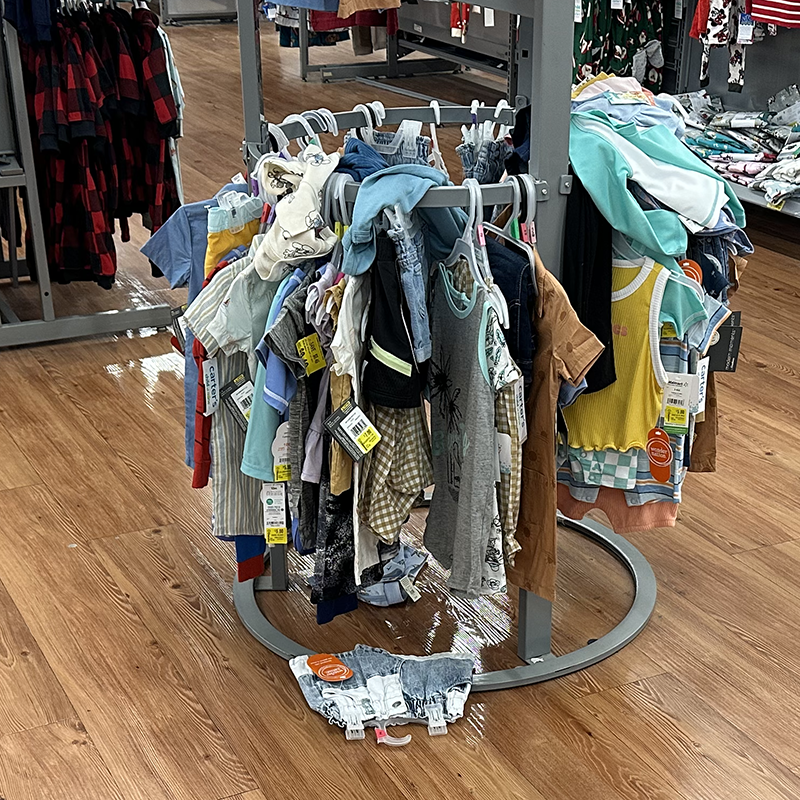 A rack of children's clothes in a department store, with some items on the floor and some careless thrown on the rack.