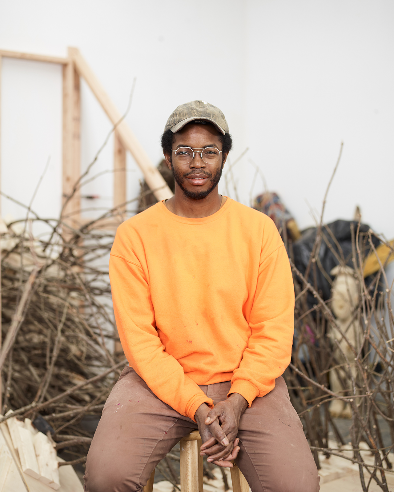A candid portrait of an artist, Hugh Hayden, seated on a wooden stool. He wears an eye-catching orange sweatshirt and brown trousers, with a camouflage cap perched atop his head. His hands are clasped in front of him, and he sports glasses with a thin frame. Behind him, a studio setting is visible, filled with an array of natural and constructed wooden forms that create a sense of creative chaos. His expression is serene and contemplative, suggesting a quiet confidence