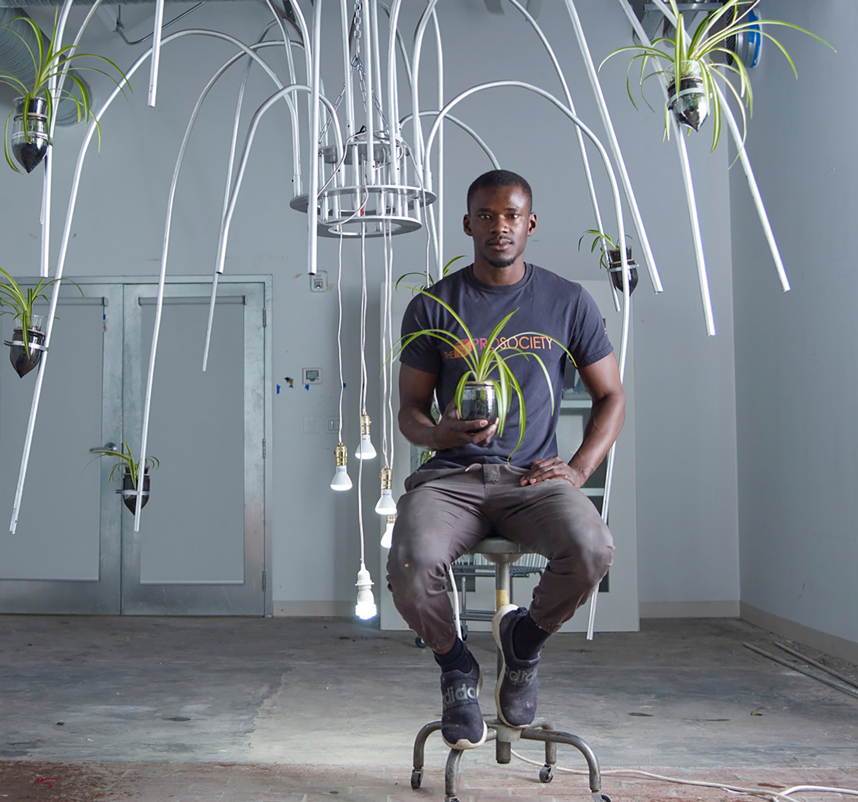 An artist sits confidently on an industrial stool in a spacious studio, surrounded by his intriguing installations. He holds a potted plant with long, green leaves that mimic the organic forms of his art pieces. Overhead, a unique chandelier of curved metal pipes and suspended light bulbs intertwines with more greenery, creating a fusion of technology and nature. His focused gaze, coupled with a casual grey t-shirt and rugged work pants, speaks to his hands-on approach to his craft. The space around him is a blend of the utilitarian and the botanical, reflective of his artistic vision.
