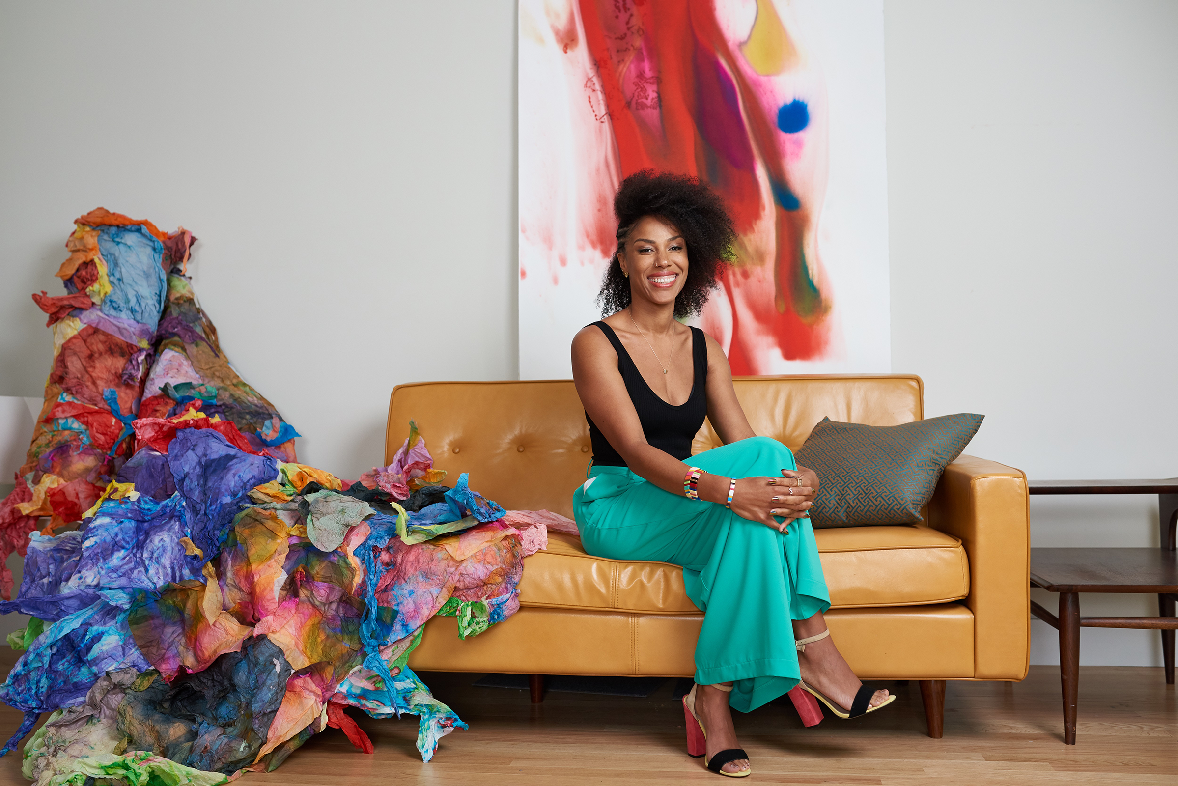 A radiant artist sits gracefully on a tan leather couch, her wide smile as engaging as the colorful art that surrounds her. She's dressed in a stylish black tank top paired with flowing teal trousers, and her red and gold heels add a pop of contrast. Her natural hair is styled in a stunning afro, and her wrists are adorned with colorful bracelets, mirroring the exuberance of the art piece. To her left, a striking sculpture made of crinkled, multicolored paper cascades onto the floor, while a large, abstract painting with swirls of red, blue, and yellow adds a dynamic backdrop to this creative scene.