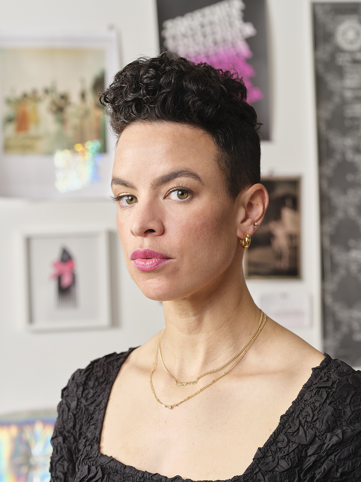 A powerful close-up of a woman artist in a gallery setting. She exudes confidence and intensity, with short, curly black hair and striking features. Her eyes are thoughtful, perhaps challenging the viewer, highlighted with makeup that accentuates her expressive gaze. She wears a textured black top that adds depth to the composition, along with tasteful gold jewelry—a pair of earrings and layered necklaces. In the background, the white walls are adorned with various artworks that speak to the vibrancy of the creative space she inhabits.