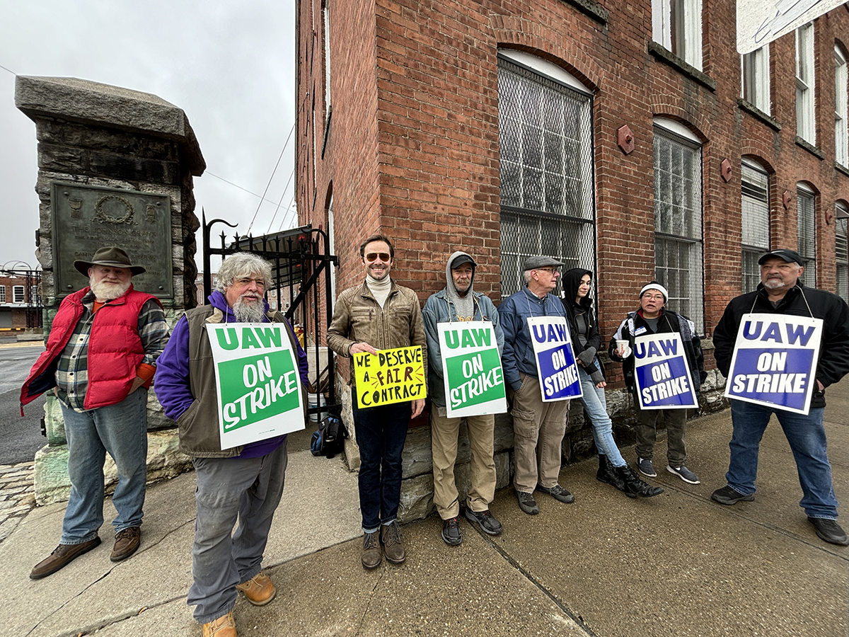 A determined group of seven MASS MoCA employees, displaying a spirit of camaraderie, stand side by side on a cloudy day. They hold a variety of signs with the UAW logo and messages like 'UAW ON STRIKE' and 'WE DESERVE A FAIR CONTRACT,' showcasing their collective bargaining efforts.