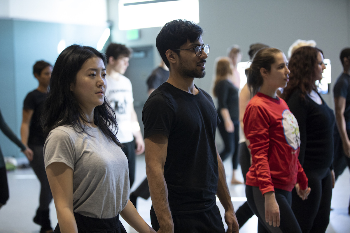 A diverse group of people practicing in a dance studio, focusing intently while standing in a line. The man in the foreground is wearing glasses and a black shirt, with the woman next to him in a gray t-shirt, both part of the workshop, "Tuning your Instrument.