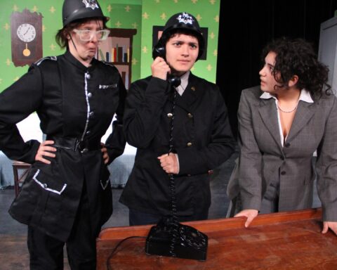 Three actors in costume during a scene from "Lights Off" by Mount Greylock Regional School. Two actors, dressed as police officers, stand beside a third actor in a suit. One officer is talking on an old-fashioned rotary phone while the other looks on with a serious expression. The background features a set designed to look like a classic British manor house.