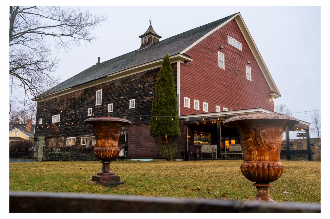 A large, rustic barn with a weathered wooden exterior and a red facade stands prominently in a grassy area. Two large, ornate rusted urns are positioned in the foreground, adding a touch of antiquity to the scene. Soft string lights are draped across the barn, and a tall evergreen tree is nestled against the side, enhancing the cozy, inviting atmosphere. The sky is overcast, contributing to the serene and picturesque setting
