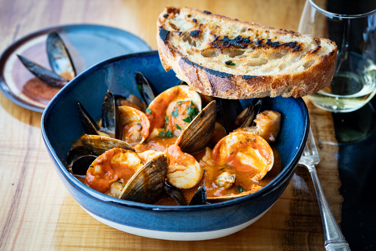 A vibrant bowl of seafood stew featuring mussels, shrimp, and clams in a rich tomato broth, garnished with fresh herbs. A slice of grilled bread rests on top of the bowl, ready for dipping. The dish is served on a wooden table with a glass of white wine and a fork nearby, creating an inviting and delicious presentation.