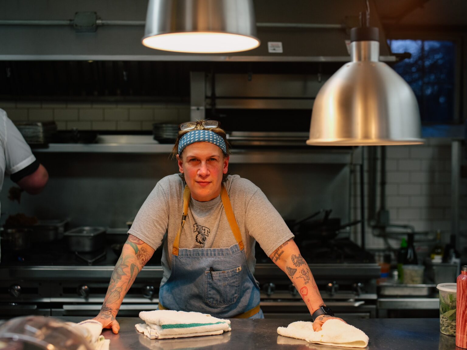 Chef Michele Ragussis, the Head Chef at Gedney Farm since last February, stands confidently in a professional kitchen. She leans on a stainless steel counter with folded kitchen towels in front of her. Wearing a gray t-shirt, a blue apron, and a blue and white bandana, she has tattoos visible on her forearms. Industrial pendant lights hang above, illuminating the space, which features stainless steel appliances and a tiled backsplash. Chef Ragussis gazes directly at the camera with a determined expression.