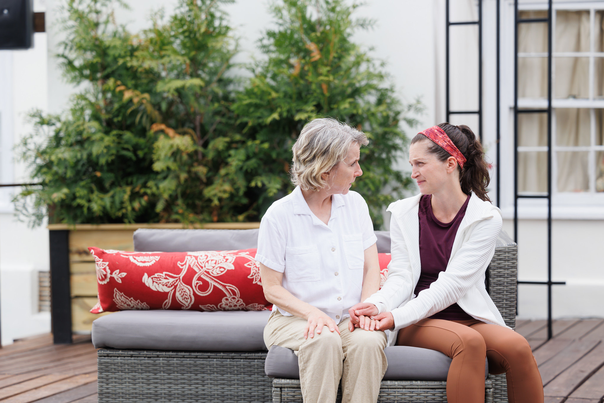 Bella Merlin, portraying Avis, and Caroline Calkins, portraying Wren in "A Body of Water," sit close together on a wicker couch with red floral cushions. Merlin, dressed in a white shirt and beige pants, looks intently at Calkins, who is wearing a maroon top, light brown leggings, and a white jacket. They hold hands and share a tender, emotional moment. The background includes green foliage and part of the house, enhancing the intimate and heartfelt atmosphere of the scene.