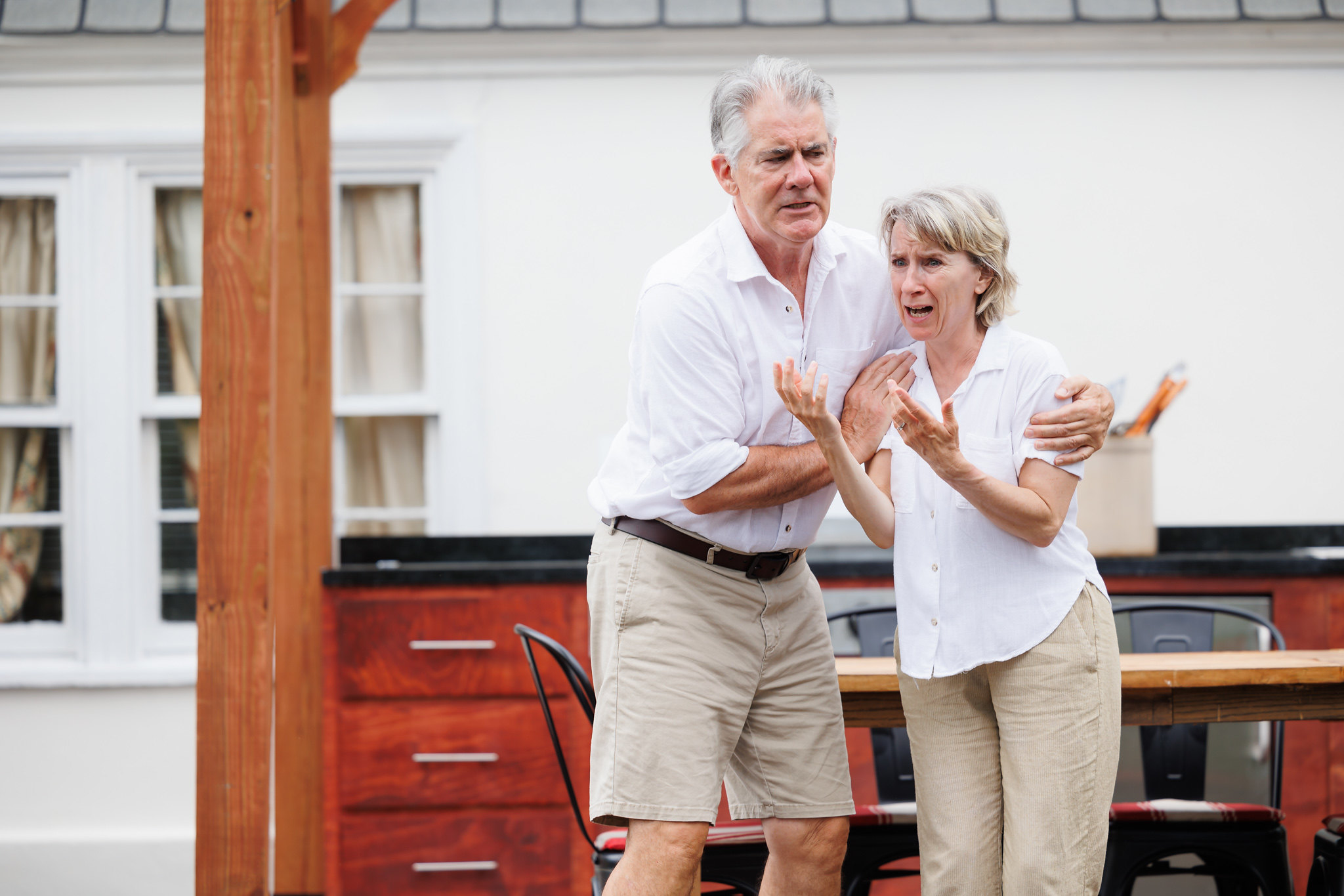 Kevin O'Rourke and Bella Merlin, portraying Moss and Avis in "A Body of Water," stand close together in a moment of intense emotion. O'Rourke has his arm around Merlin, who appears distressed and is gesturing with her hands. Both are dressed in white shirts and beige shorts. The background features part of the house with a window and an outdoor kitchen area, emphasizing the domestic yet unsettling setting of the scene.