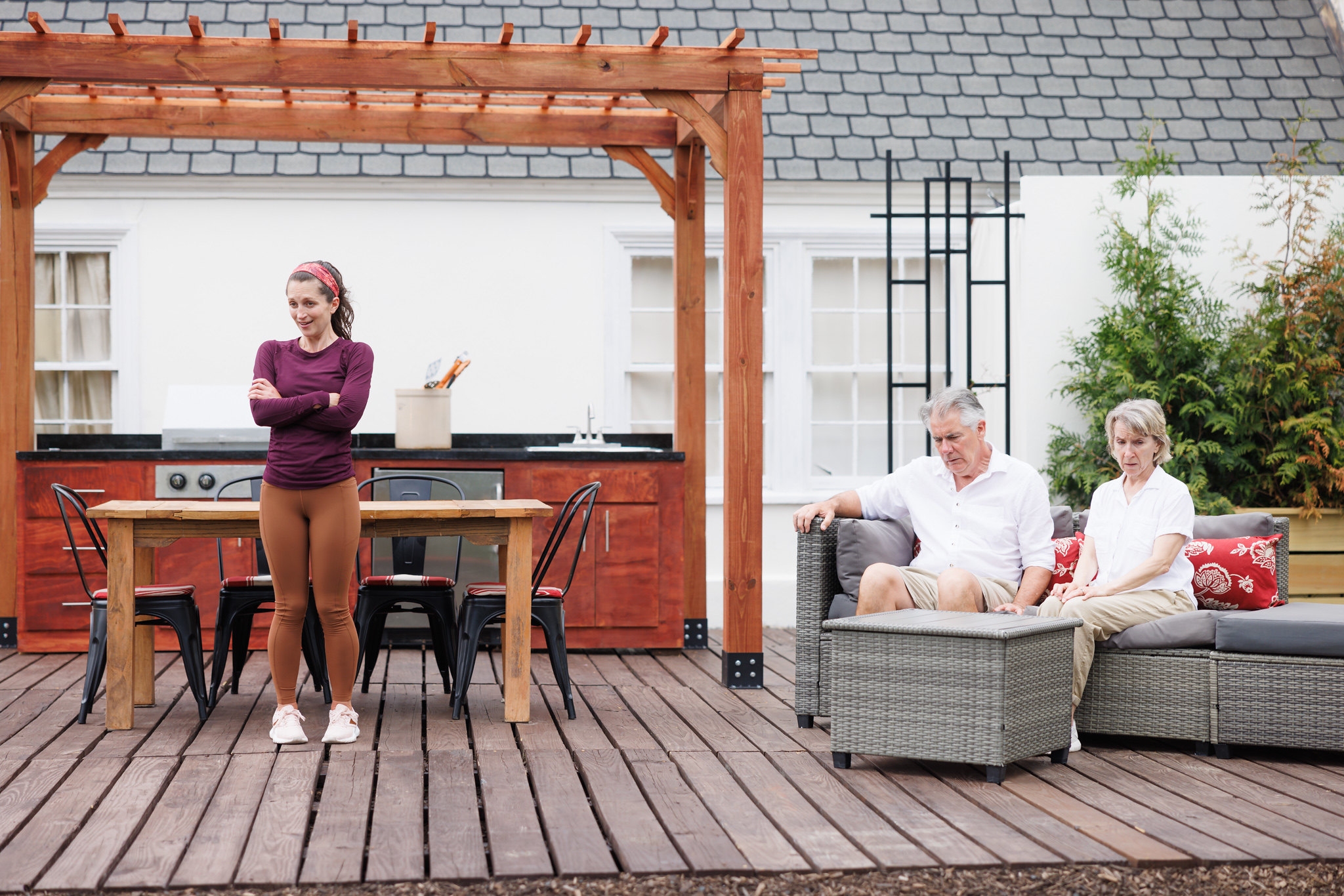 Alt text: Caroline Calkins, as Wren in "A Body of Water," stands with her arms crossed and a thoughtful expression in front of a wooden table on an outdoor deck. She is dressed in a long-sleeved maroon top and light brown leggings. To the right, Kevin O'Rourke and Bella Merlin, portraying Moss and Avis, sit on a wicker couch with red floral cushions. They both wear white shirts and beige shorts, looking down contemplatively. The background shows part of the house, an outdoor kitchen area, and greenery, suggesting a serene yet tense scene from the play.