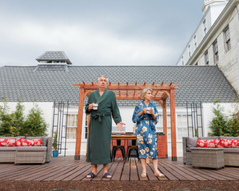 Kevin O'Rourke and Bella Merlin, who portray Moss and Avis in Lee Blessing's "A Body of Water," stand on a wooden deck in front of a well-appointed house. O'Rourke is in a green robe and slippers, holding a cup of coffee, and gesturing with one hand, while Merlin, in a blue floral robe and sandals, also holds a coffee cup. They appear contemplative and somewhat confused, fitting the theme of the play. The background shows part of the house with a trellis and outdoor seating area.