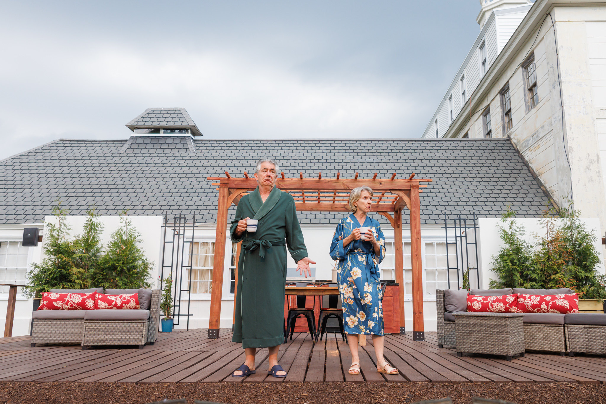 Kevin O'Rourke and Bella Merlin, who portray Moss and Avis in Lee Blessing's "A Body of Water," stand on a wooden deck in front of a well-appointed house. O'Rourke is in a green robe and slippers, holding a cup of coffee, and gesturing with one hand, while Merlin, in a blue floral robe and sandals, also holds a coffee cup. They appear contemplative and somewhat confused, fitting the theme of the play. The background shows part of the house with a trellis and outdoor seating area.
