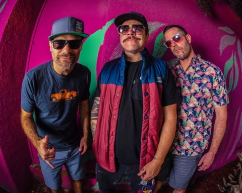 Three members of the band 'Badfish' pose against a vibrant, colorful wall with pink and green graffiti. The man on the left wears a blue T-shirt with an orange truck graphic, sunglasses, and a gray cap. The middle man sports a mustache, sunglasses, a black shirt, and a red and blue vest. The man on the right is dressed in a floral print shirt, sunglasses, and striped shorts. All three have a casual, relaxed demeanor.