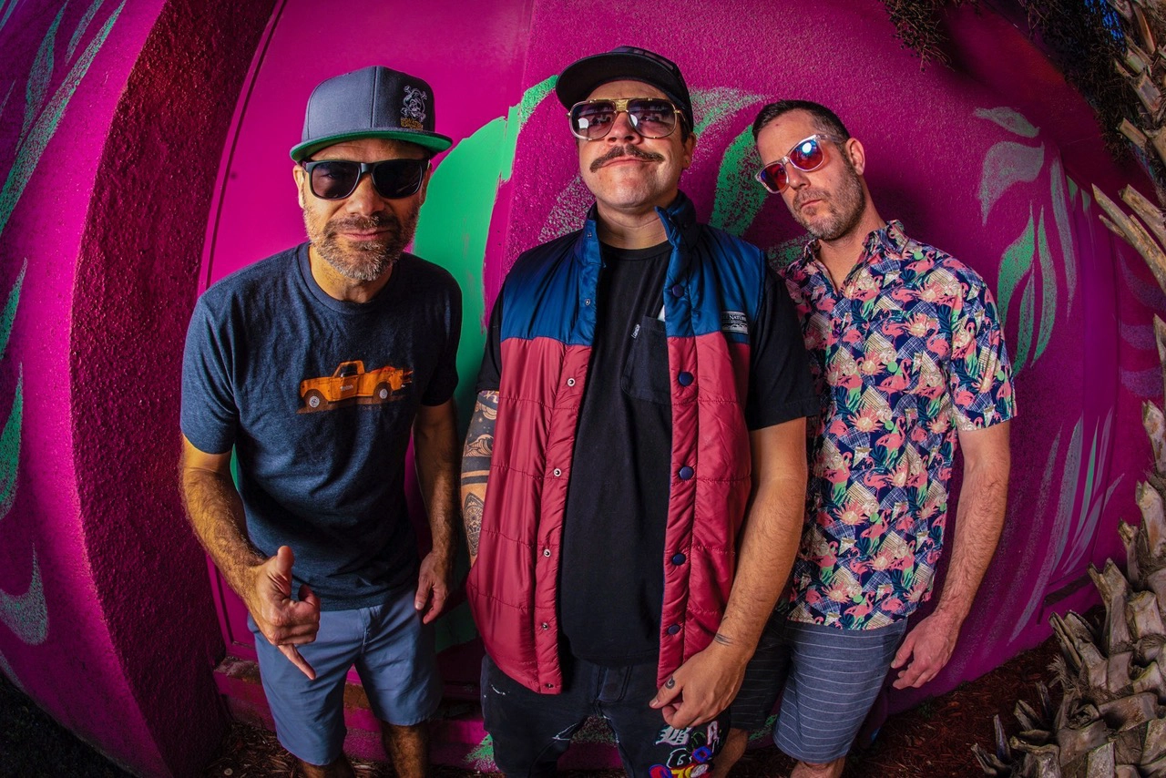 Three members of the band 'Badfish' pose against a vibrant, colorful wall with pink and green graffiti. The man on the left wears a blue T-shirt with an orange truck graphic, sunglasses, and a gray cap. The middle man sports a mustache, sunglasses, a black shirt, and a red and blue vest. The man on the right is dressed in a floral print shirt, sunglasses, and striped shorts. All three have a casual, relaxed demeanor.