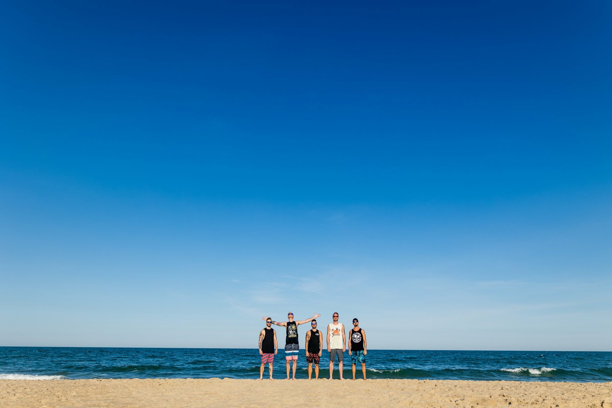 Five members of the band 'Ballyhoo' stand on a sandy beach with the ocean in the background under a clear blue sky. Each member is dressed in casual summer attire, including tank tops and shorts. The central figure has their arms outstretched, while the others stand relaxed with their hands at their sides.