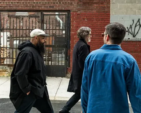 Three men walking along a city sidewalk with brick buildings in the background. The man on the left wears a black jacket and white cap, the man in the middle has long hair and a black coat, and the man on the right wears glasses and a blue jacket.