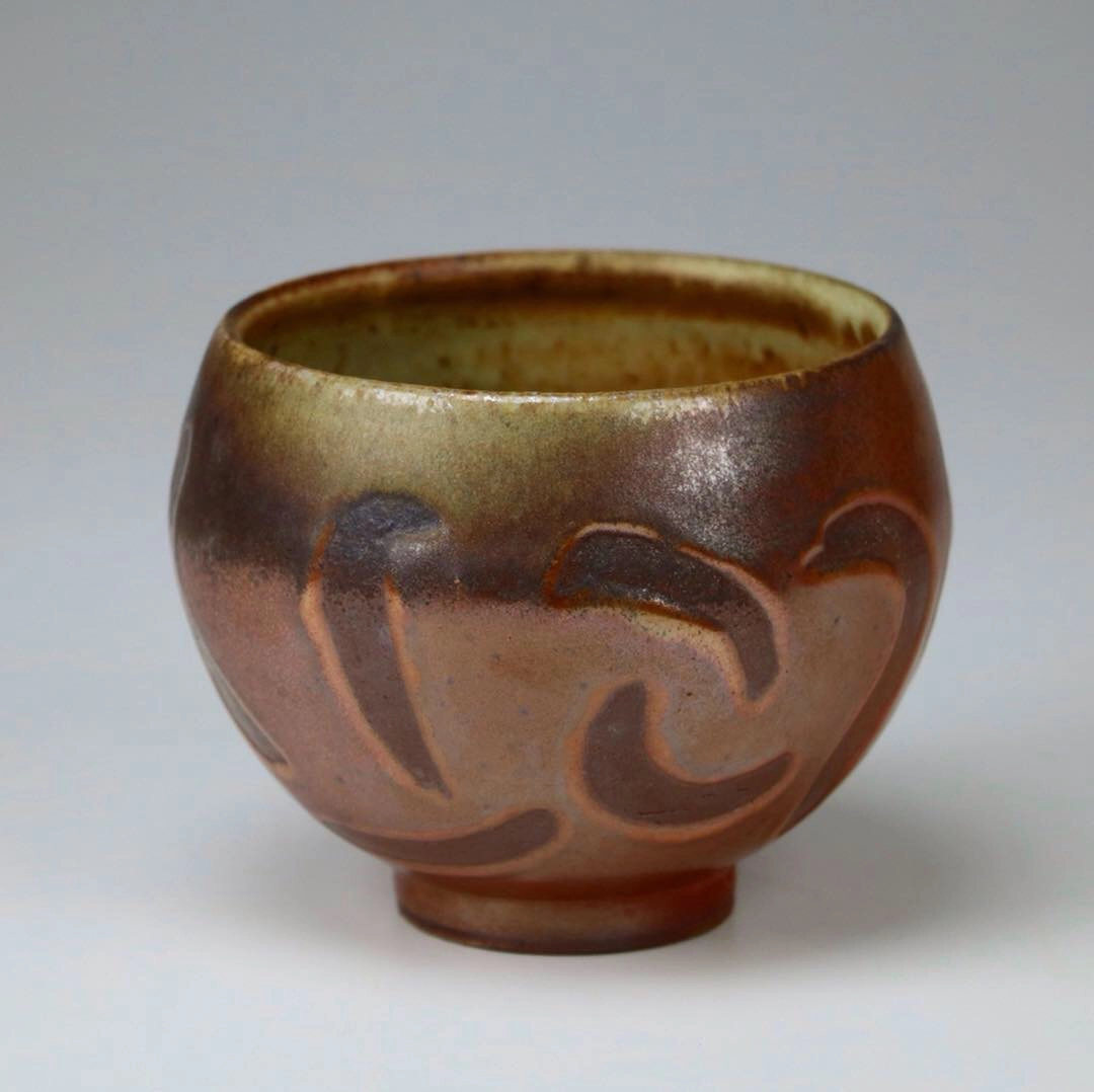 A handcrafted ceramic bowl by Hunter Cady. The bowl features a rounded shape with a slightly flared rim and a raised foot. Its surface is adorned with earthy brown and green glazes, creating a natural, rustic appearance. The exterior is decorated with subtle, flowing carved patterns, adding texture and visual interest to the piece.