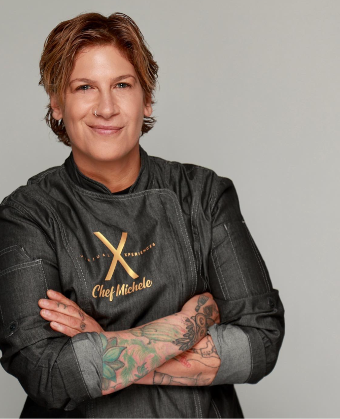 Chef Michele Ragussis stands confidently with arms crossed, wearing a dark chef's coat with the name 'Chef Michele' embroidered in gold. She has short, tousled hair and tattoos on her forearms, showcasing a blend of professional and personal style. The background is a simple gray, drawing focus to her friendly and determined expression.