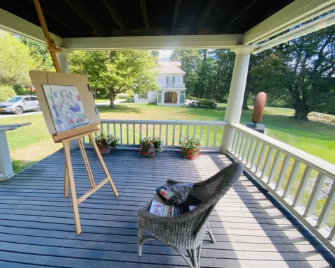 A scenic porch setup for figure drawing at Chesterwood. An easel with a drawing of a seated figure is positioned near the edge of the porch, overlooking a lush, green lawn with trees and a historic white house in the background. A wicker chair with art supplies sits nearby. Potted flowers decorate the porch railing, adding a touch of color to the serene and picturesque setting.