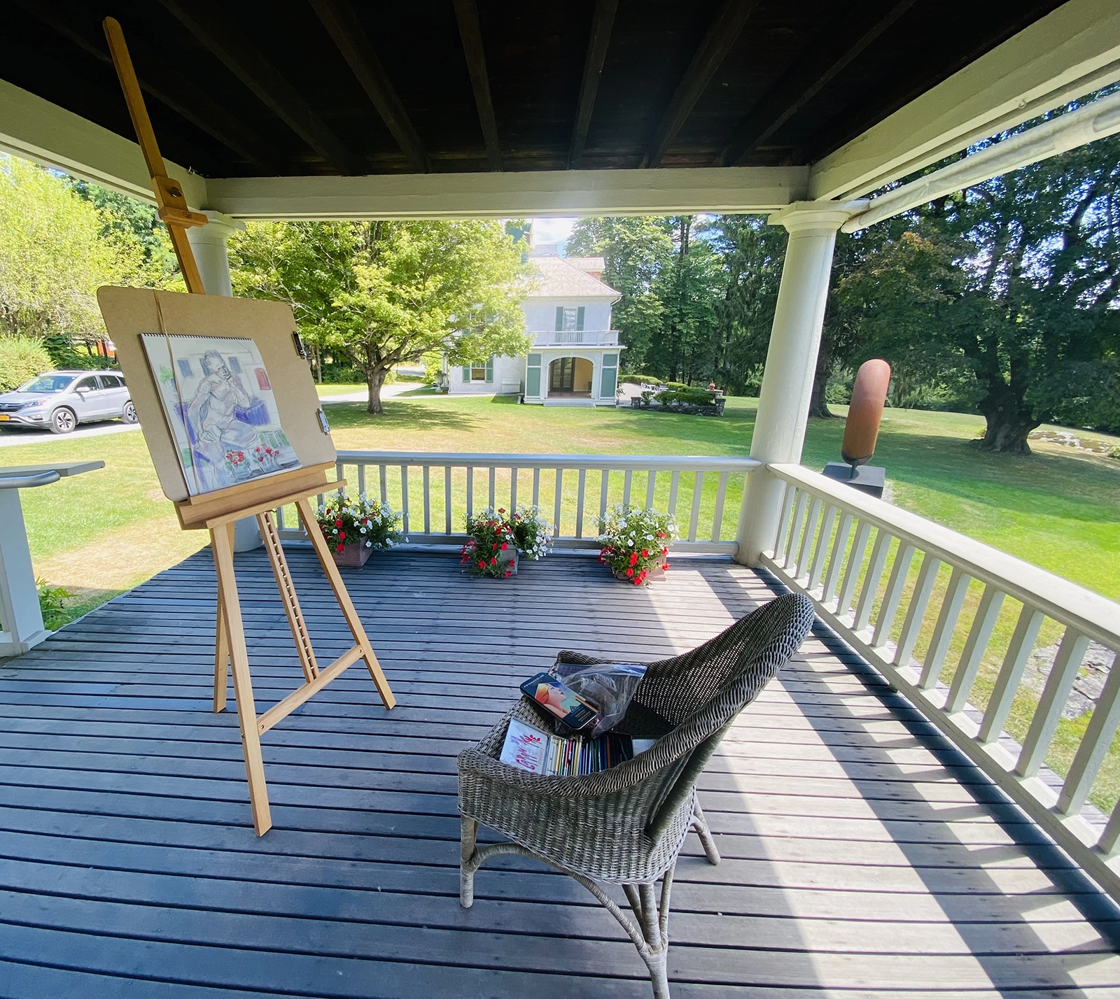 A scenic porch setup for figure drawing at Chesterwood. An easel with a drawing of a seated figure is positioned near the edge of the porch, overlooking a lush, green lawn with trees and a historic white house in the background. A wicker chair with art supplies sits nearby. Potted flowers decorate the porch railing, adding a touch of color to the serene and picturesque setting.