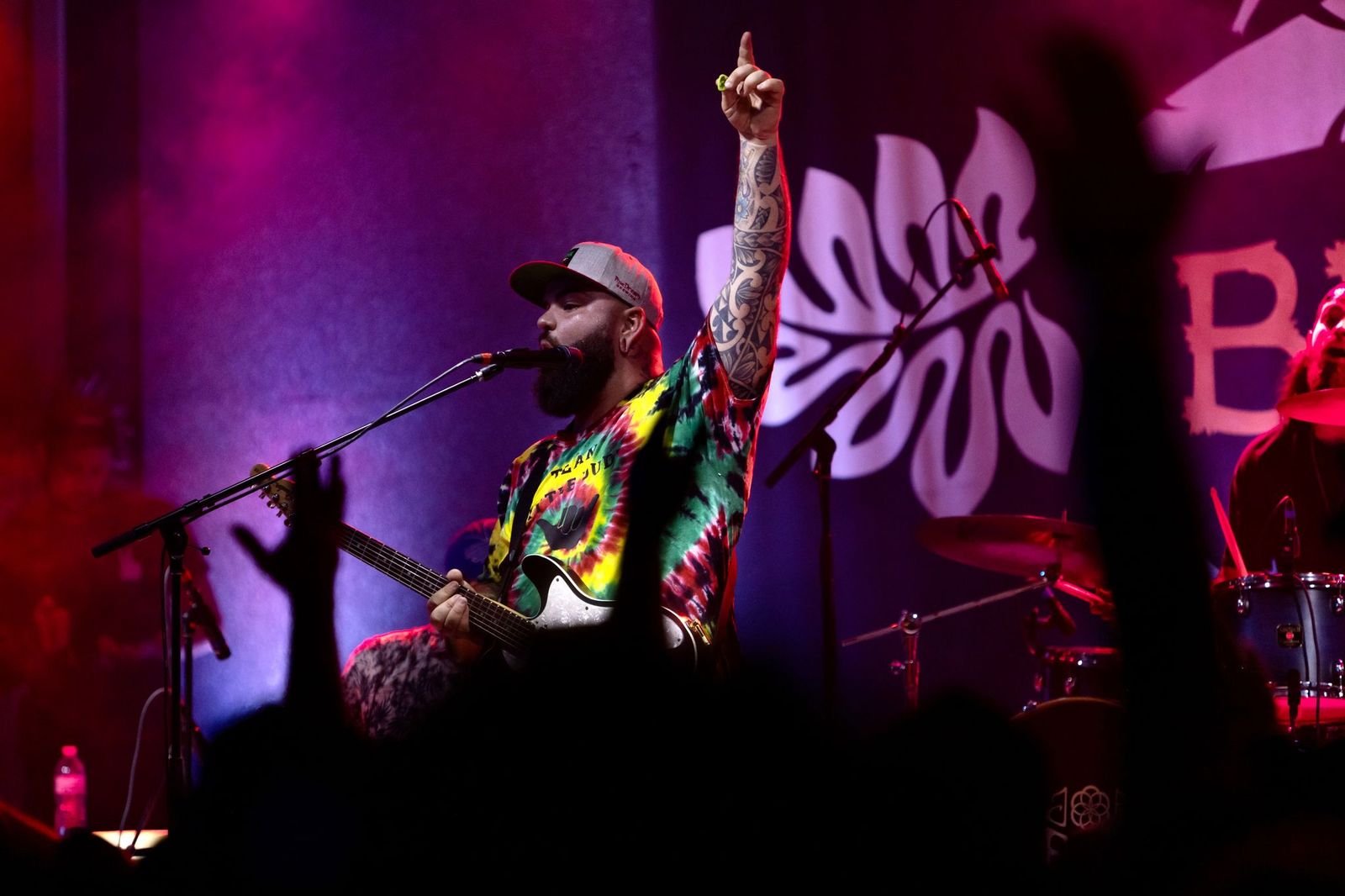 Music artist Joe Samba performs on stage, playing a guitar and singing into a microphone. He is wearing a colorful tie-dye shirt and a gray cap, with his tattooed arm raised and pointing upwards. The background features a vibrant stage setup with purple and pink lighting and a drum set. The silhouettes of the audience's raised hands are visible in the foreground, adding to the lively concert atmosphere.