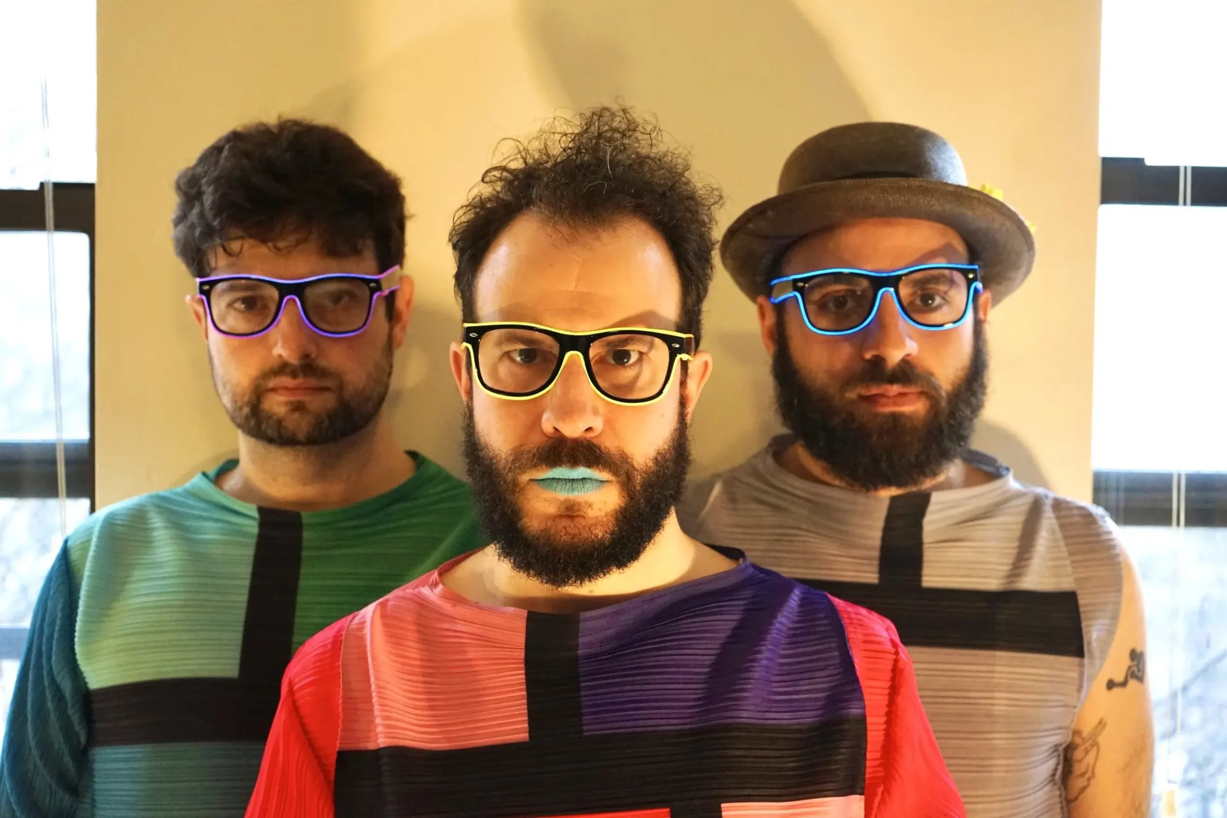 Three male members of the band Pinc Louds stand in a row, wearing colorful geometric-patterned shirts and neon glasses, with the lead singer sporting blue lipstick, against a plain background.