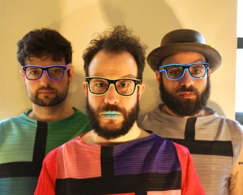 Three male members of the band Pinc Louds stand in a row, wearing colorful geometric-patterned shirts and neon glasses, with the lead singer sporting blue lipstick, against a plain background.
