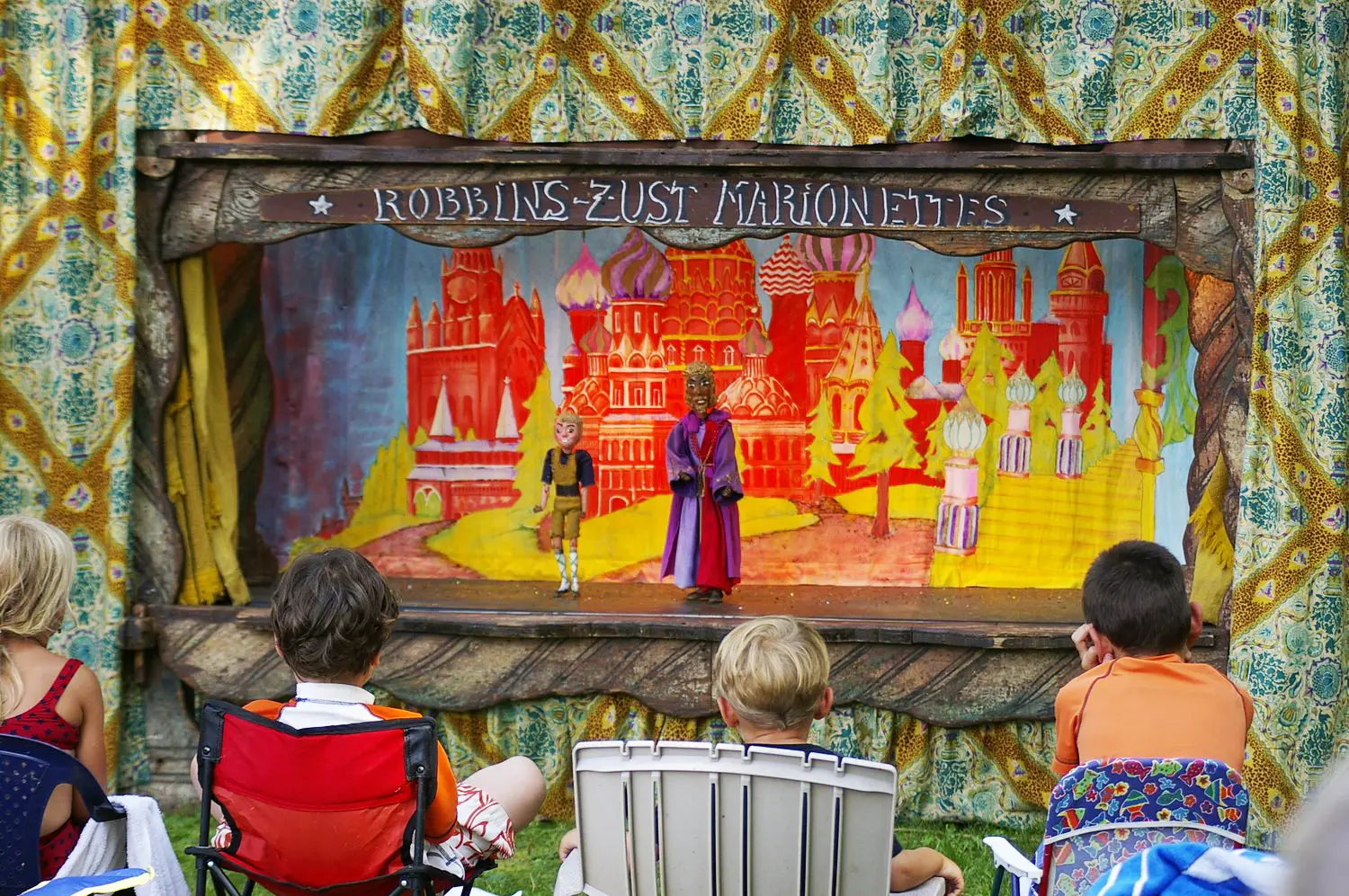 Children watching a colorful marionette performance by Robbins-Zust Marionettes, featuring two puppets on stage against a backdrop of vibrant, painted scenery.