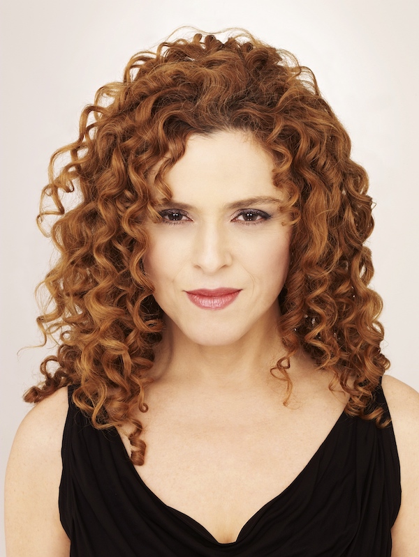 A headshot of a woman, Bernadette Peters, featuring her signature curly red hair cascading around her face. She is wearing a black top and has a subtle, confident smile with a direct, engaging gaze. The background is a soft, neutral tone, highlighting her vibrant hair and expressive features.