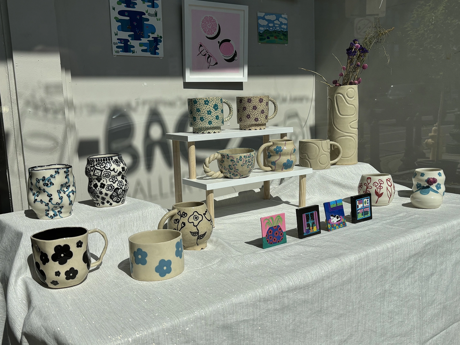 A display of handcrafted pottery by Abby Klein, featuring a variety of mugs and vases adorned with whimsical and intricate patterns. The pottery pieces are arranged on a white cloth-covered table, with some elevated on a small wooden shelf. The designs include blue and black floral motifs, red abstract patterns, and other decorative elements. Small framed artworks and a vase with dried flowers add to the artistic presentation. Sunlight casts shadows and reflections, enhancing the vibrant and creative atmosphere of the display.