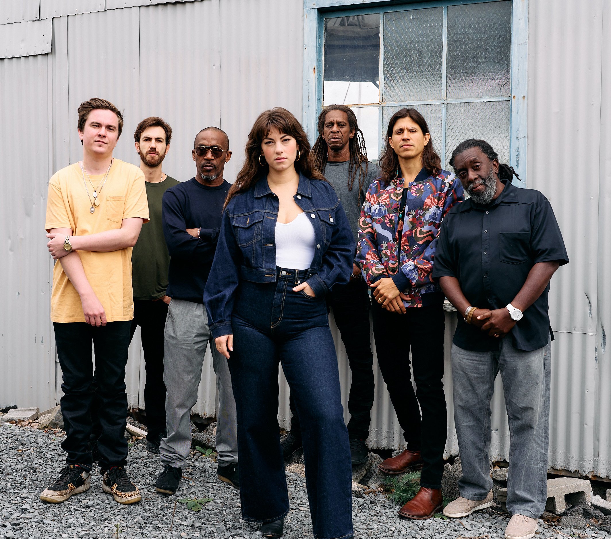 Seven members of the band 'SunDub' stand in front of a corrugated metal wall with a window. The central figure is a woman in a denim jacket and jeans, with long brown hair and hoop earrings. The men around her are dressed in a variety of casual and semi-formal outfits, including a yellow T-shirt, a patterned jacket, and dark sunglasses. The group's serious expressions add to the strong, cohesive vibe of the photo.