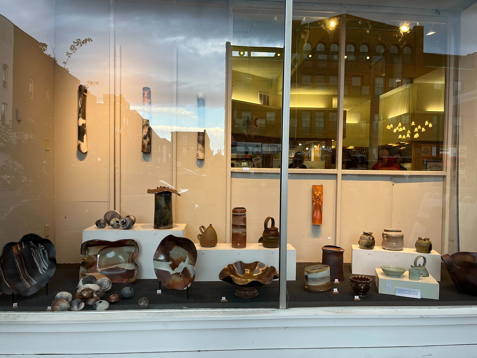 A window display of a pottery gallery showcasing various ceramic pieces. The display includes a variety of vases, bowls, and decorative objects arranged on white pedestals and shelves. The pottery features different shapes, sizes, and earth-toned glazes. Reflections of buildings and the sky can be seen on the window glass, adding depth to the scene. Inside the gallery, warm lighting illuminates the artworks, creating an inviting atmosphere.