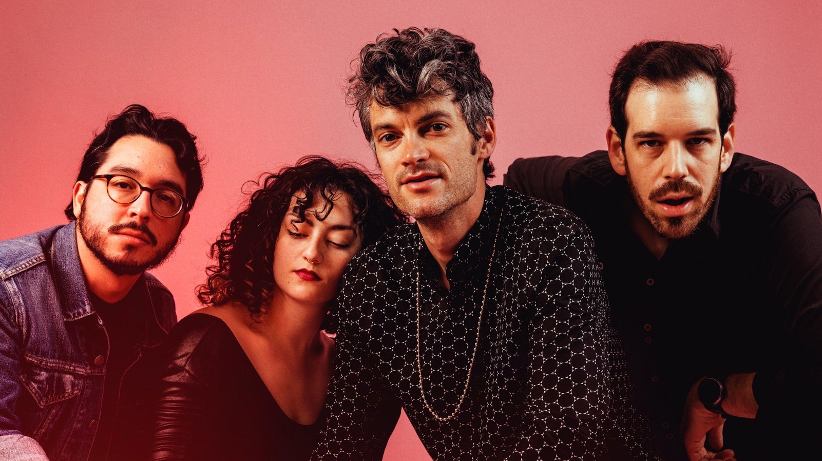 A group photo of the band Billy Wylder. The image features four members against a pink background. On the left, a man with short dark hair, glasses, and a beard wears a denim jacket. Next to him, a woman with curly dark hair and a red lipstick has her eyes closed and wears a black top. The third member, with wavy, graying hair and a patterned black shirt, looks directly at the camera with a slight smile. The fourth member, on the far right, has short dark hair and a beard, wearing a black shirt and looking intently at the camera.