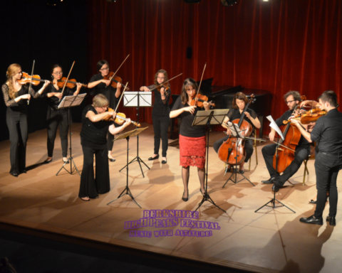 A group of musicians performs on stage, each playing a string instrument, including violins and cellos. They are dressed in formal attire with a red curtain backdrop. The ensemble includes both male and female performers, and they are positioned in a semi-circle. The text "Berkshire High Peaks Festival: Music with Altitude!" is visible at the bottom of the image.