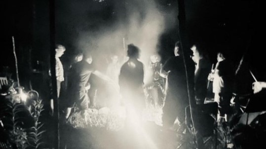 A group of people gather in a circle around a blazing fire in a dark, forested area. The scene is illuminated by the firelight, casting shadows and creating a mystical, eerie atmosphere. The figures are mostly in silhouette, with some details of their clothing and faces visible. Smoke rises from the fire, adding to the ethereal quality of the image. The photo captures a sense of ritual or celebration in a natural, nocturnal setting.