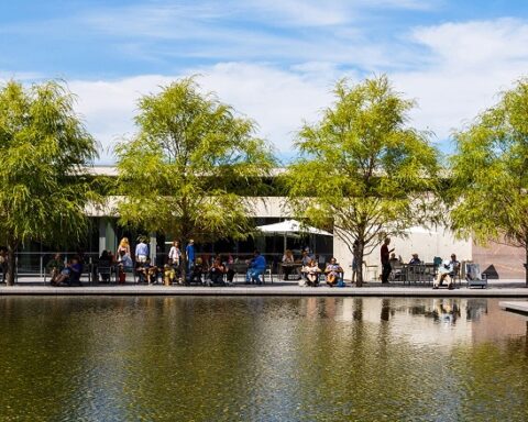 "A view of the Clark Art Institute and its reflecting pool on a sunny day. The scene features a modern building with a clean, minimalist design, lined with tall, leafy trees. People are seated and walking along the pathway by the water, enjoying the serene atmosphere. The reflecting pools mirror the blue sky and surrounding greenery. Photo by Tucker Bair."