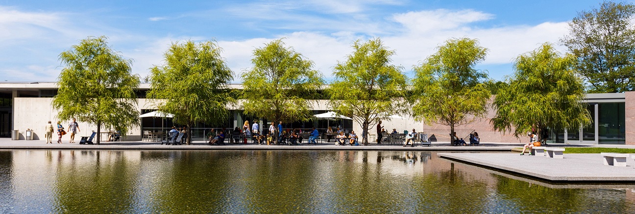 "A view of the Clark Art Institute and its reflecting pool on a sunny day. The scene features a modern building with a clean, minimalist design, lined with tall, leafy trees. People are seated and walking along the pathway by the water, enjoying the serene atmosphere. The reflecting pools mirror the blue sky and surrounding greenery. Photo by Tucker Bair."