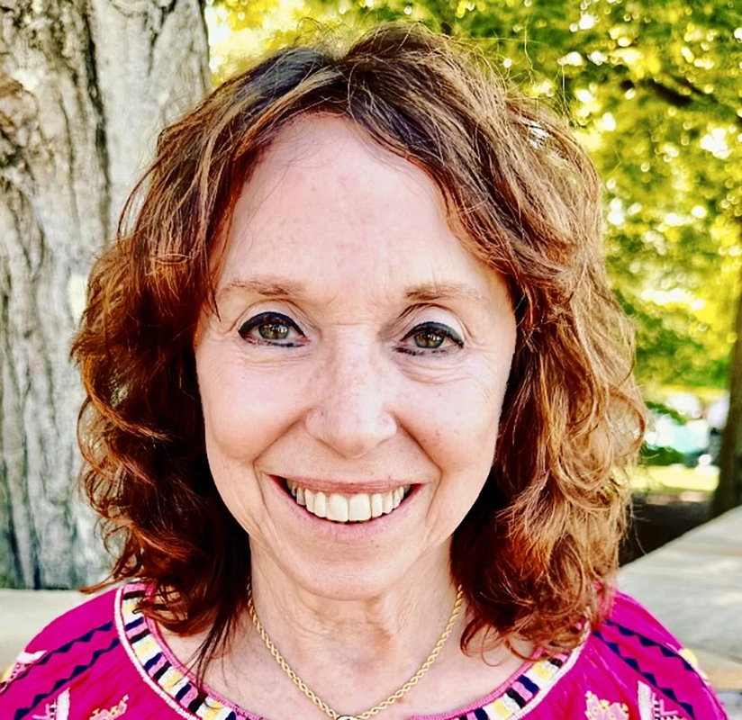 Headshot of a woman, Alison Bendix, playwright of "The Refuseniks." Alison is smiling, with curly, reddish-brown hair and is wearing a pink top with colorful embroidery. The background features trees and greenery, suggesting an outdoor setting.