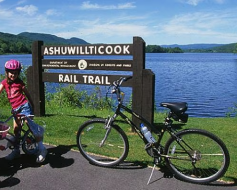 A family with bicycles enjoying a sunny day at the Ashuwillticook Rail Trail in Massachusetts. The trail sign stands near a calm, scenic lake surrounded by lush greenery and distant hills.