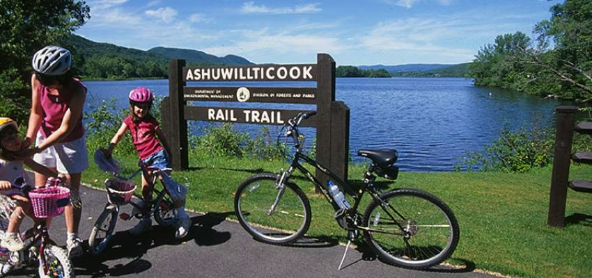 A family with bicycles enjoying a sunny day at the Ashuwillticook Rail Trail in Massachusetts. The trail sign stands near a calm, scenic lake surrounded by lush greenery and distant hills.