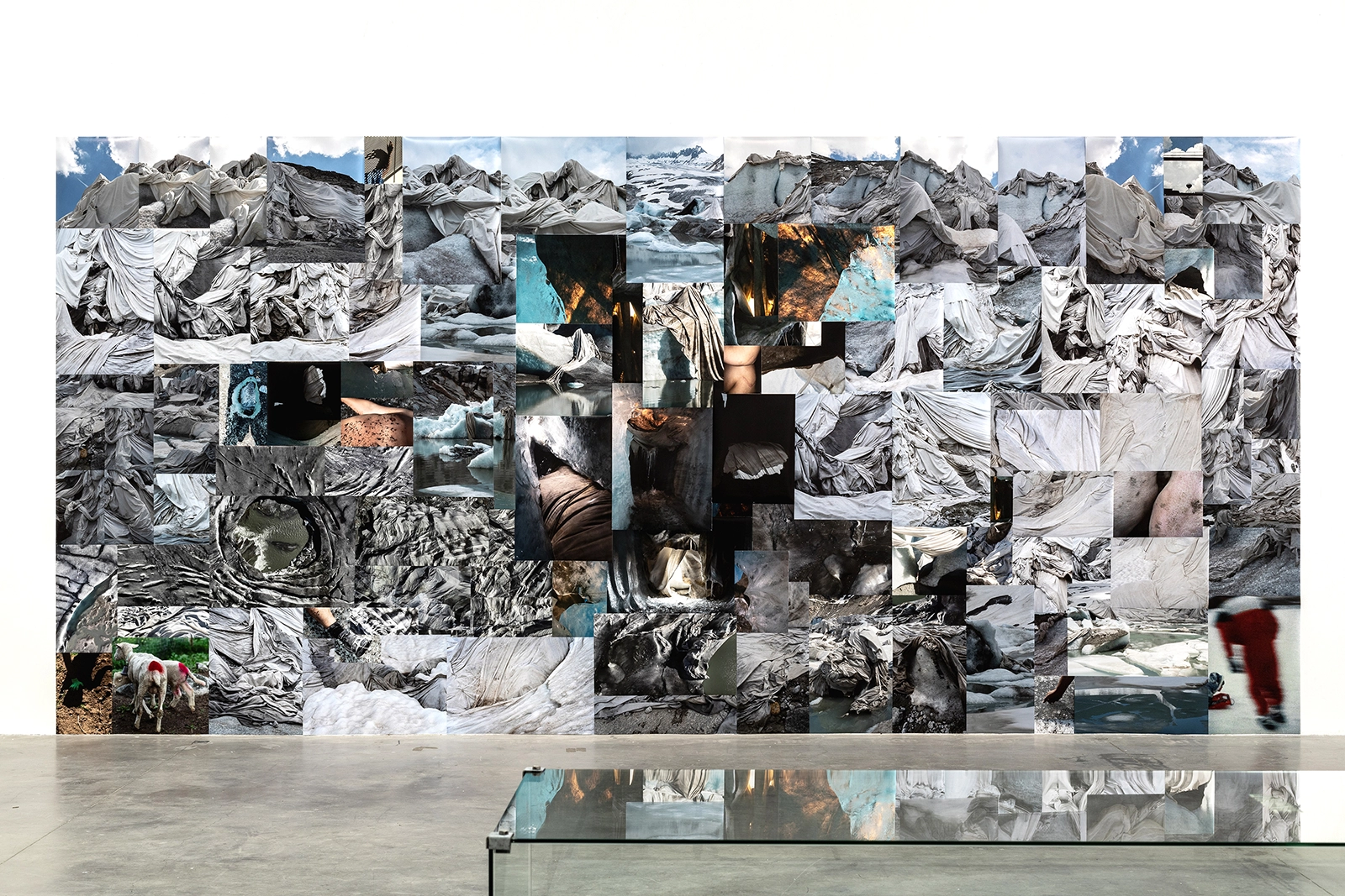 Installation view of To dress a wound from the light that shines from it (Belly of a Glacier) by Ohan Breiding. The artwork features 111 giclée prints arranged as a large collage of images depicting glaciers draped with thermal blankets, melting ice, and environmental textures. A reflective glass table is visible in the foreground, emphasizing the immersive scale of the installation.