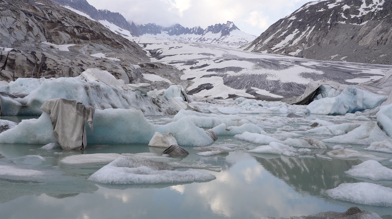 Film still from Belly of a Glacier by Ohan Breiding, showing the Rhône Glacier in Switzerland covered with thermal blankets. Ice formations and melting pools of turquoise water reflect the surrounding snow-covered mountains under a cloudy sky. The image highlights the glacier’s fragility and the impact of climate change.