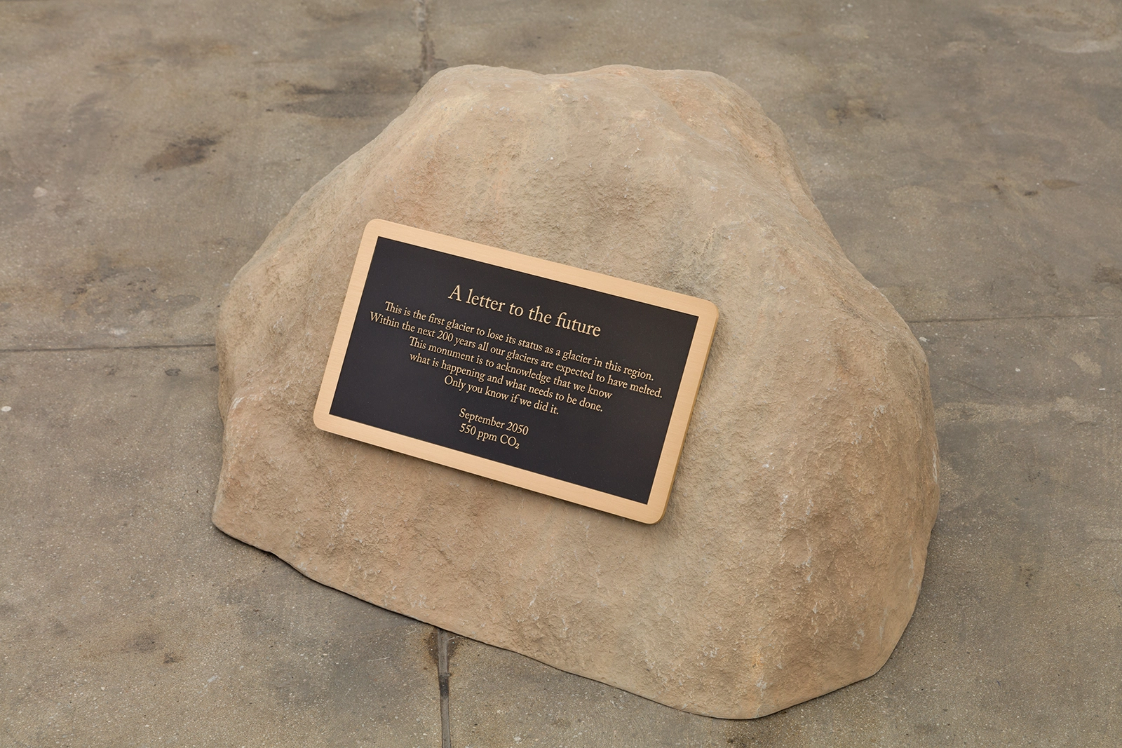 A stone monument featuring a plaque titled A letter to the future. The plaque reads: "This is the first glacier to lose its status as a glacier in this region. Within the next 200 years, all our glaciers are expected to have melted. This monument is to acknowledge that we know what is happening and what needs to be done. Only you know if we did it. September 2050, 550 ppm CO₂." The stone rests on a concrete floor.