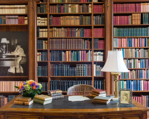 The Library at The Mount, Edith Wharton’s home, featuring floor-to-ceiling wooden bookshelves filled with colorful books. A vintage desk holds stacks of books, a bouquet of flowers, a lamp, and a framed photograph. A black-and-white portrait of Edith Wharton is displayed on the left wall.